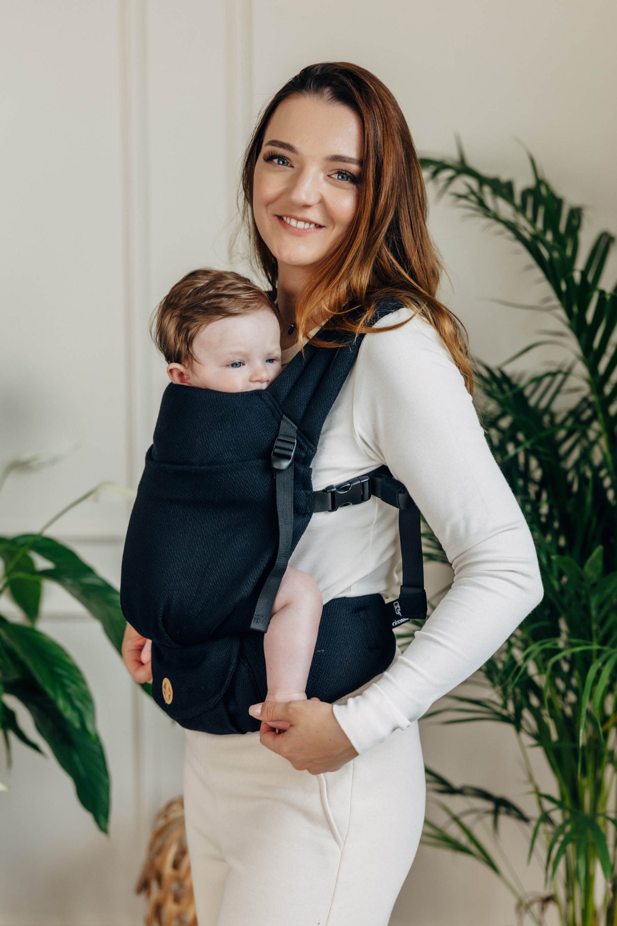 Mom wearing LennyLight black baby carrier with infant, showcasing comfortable front carrying option amidst greenery.