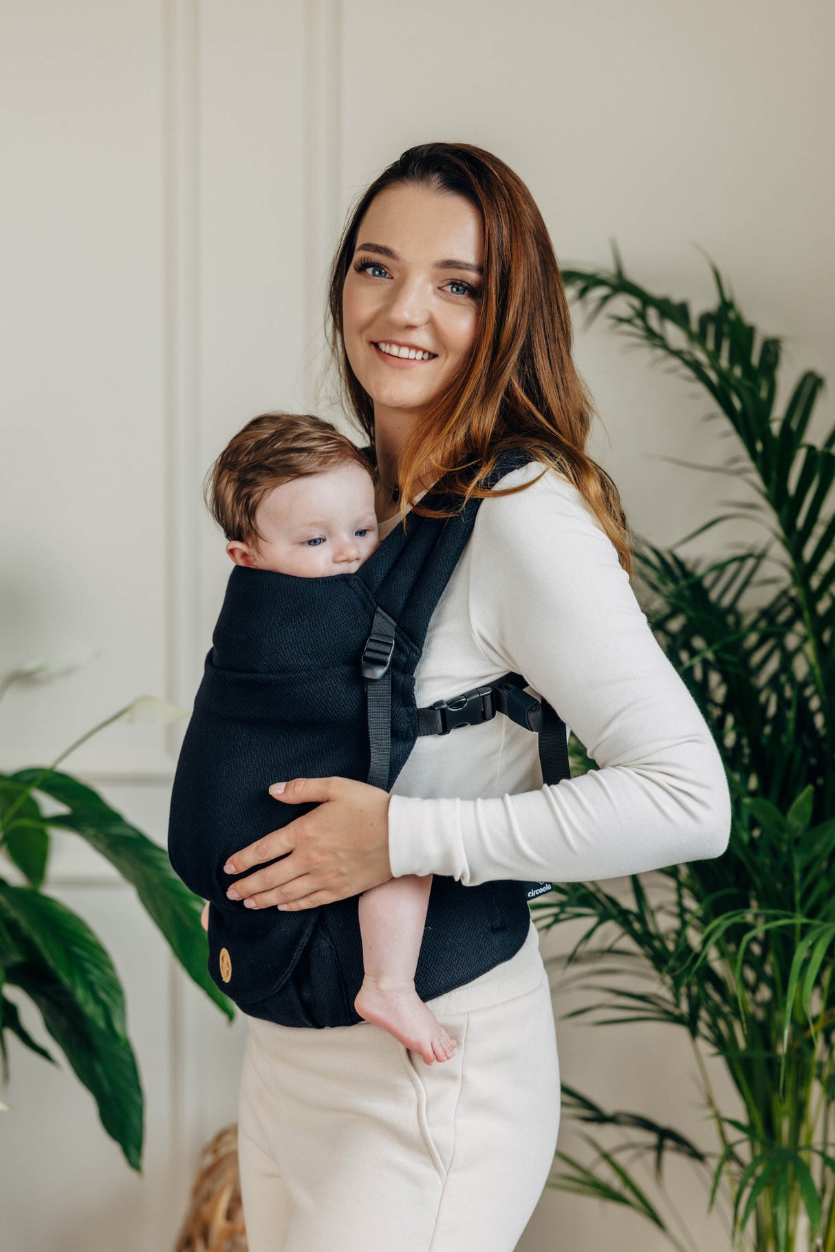 Mom wearing the black cotton LennyLight baby carrier with a baby in front against a bright indoor setting.