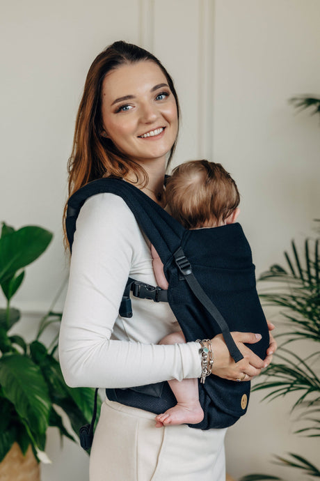 Smiling woman holding a baby in a black cotton baby carrier, LennyLight, showcasing comfort and versatility.