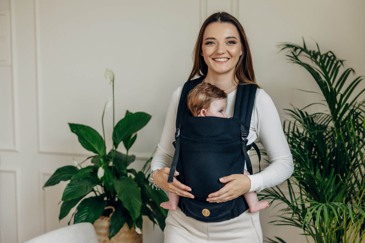 Smiling woman holding a baby in a black cotton LennyLight baby carrier against a bright indoor setting.