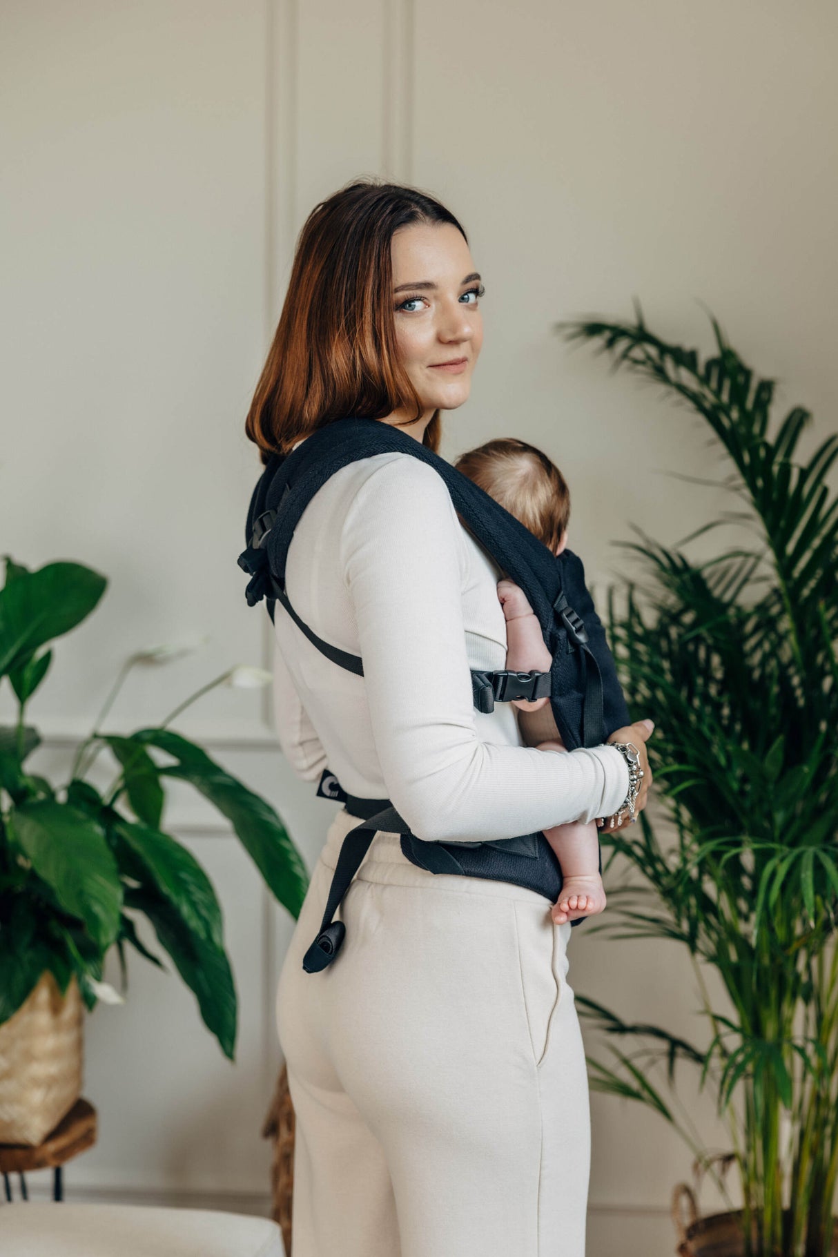 Woman wearing black cotton LennyLight baby carrier with a baby, surrounded by greenery indoors.