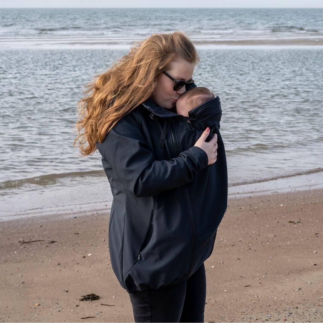 Mamalila Softshell Babywearing Jacket Allrounder - Black featuring a parent holding a baby by the beach | Babymaxi