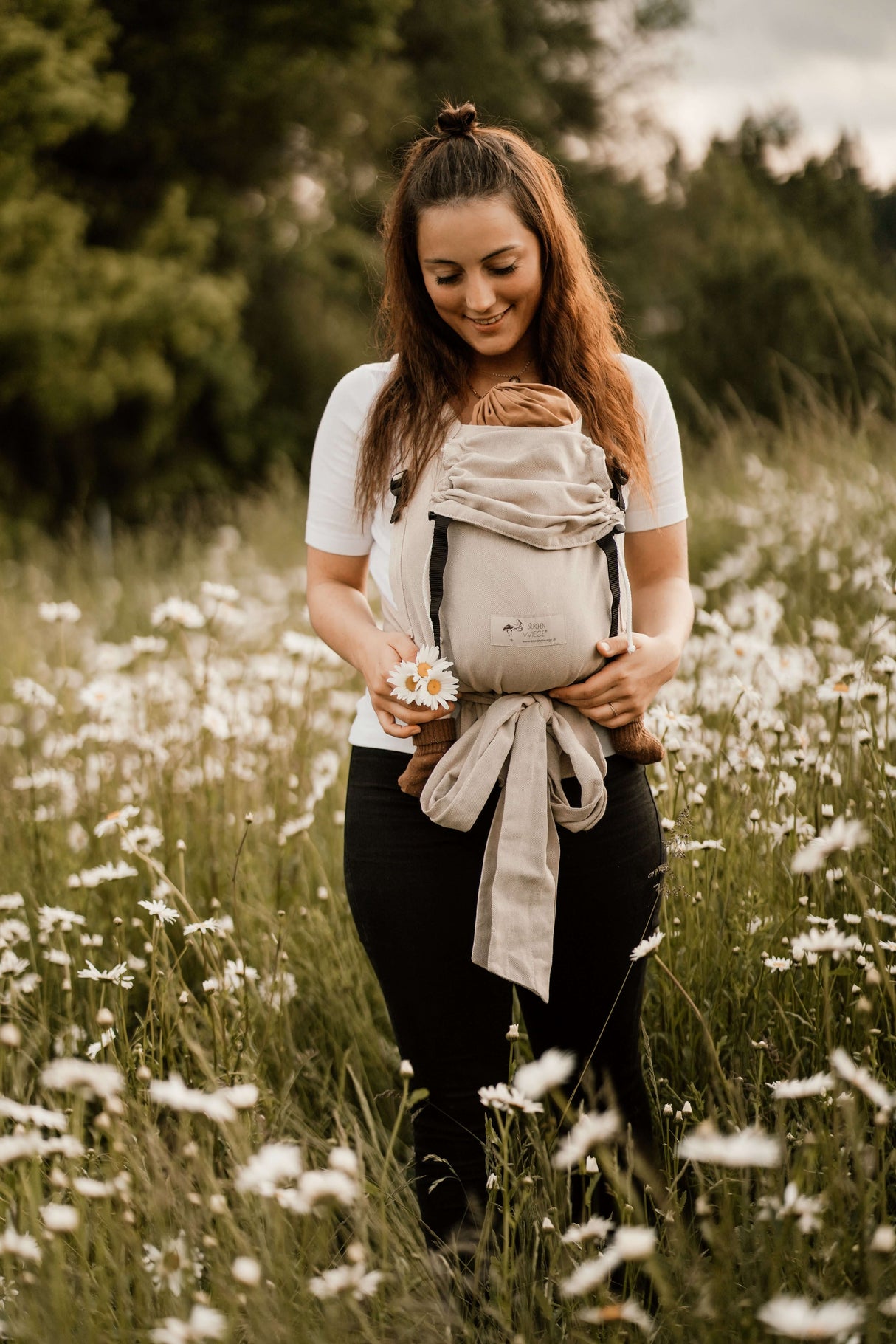 Woman standing in a field holding a baby in a Storchenwiege Talemo Half-Buckle Baby Carrier - Natural | Babymaxi