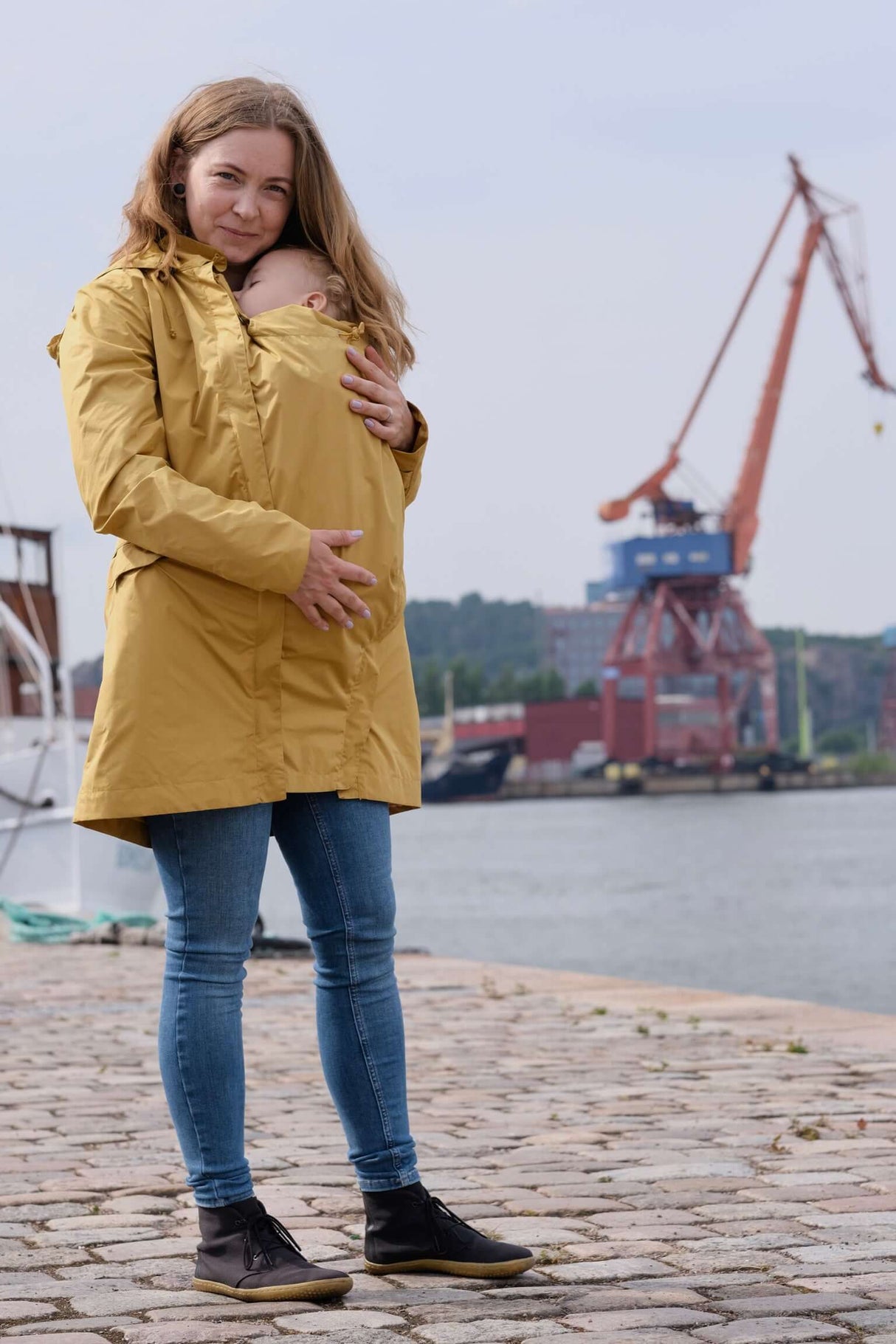 Mamalila Dublin Babywearing Coat – Mustard showcased by a woman carrying a baby, perfect for spring, summer, and autumn | Babymaxi