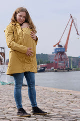 Mamalila Dublin Babywearing Coat – Mustard showcased by a woman carrying a baby, perfect for spring, summer, and autumn | Babymaxi