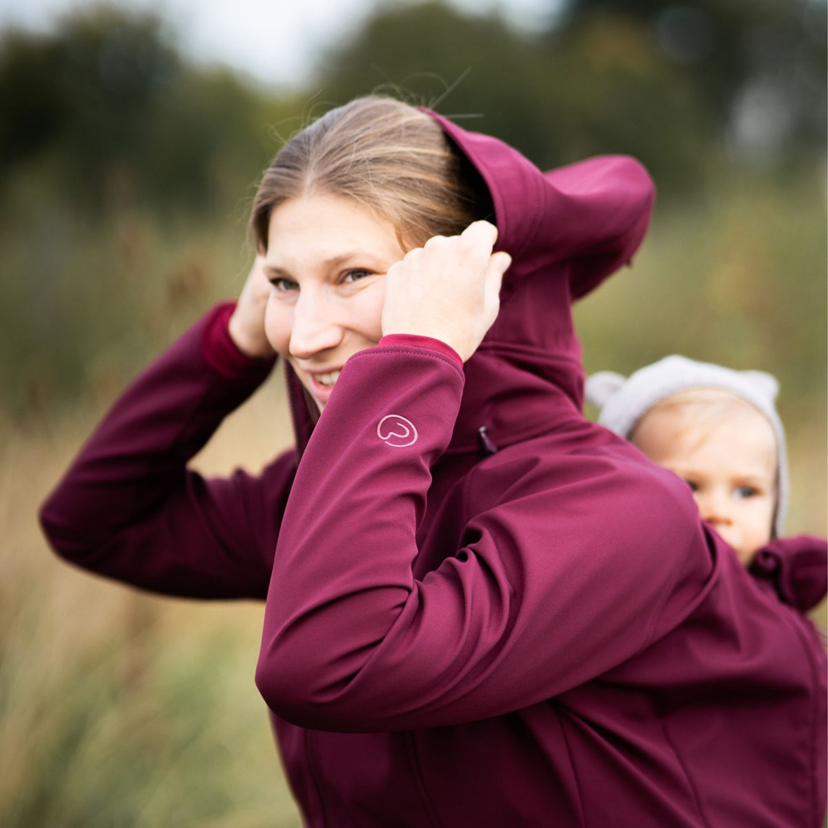 Mamalila Softshell Babywearing Jacket Allrounder Fit Burgundy, mom with baby in back carry pulling up the hood