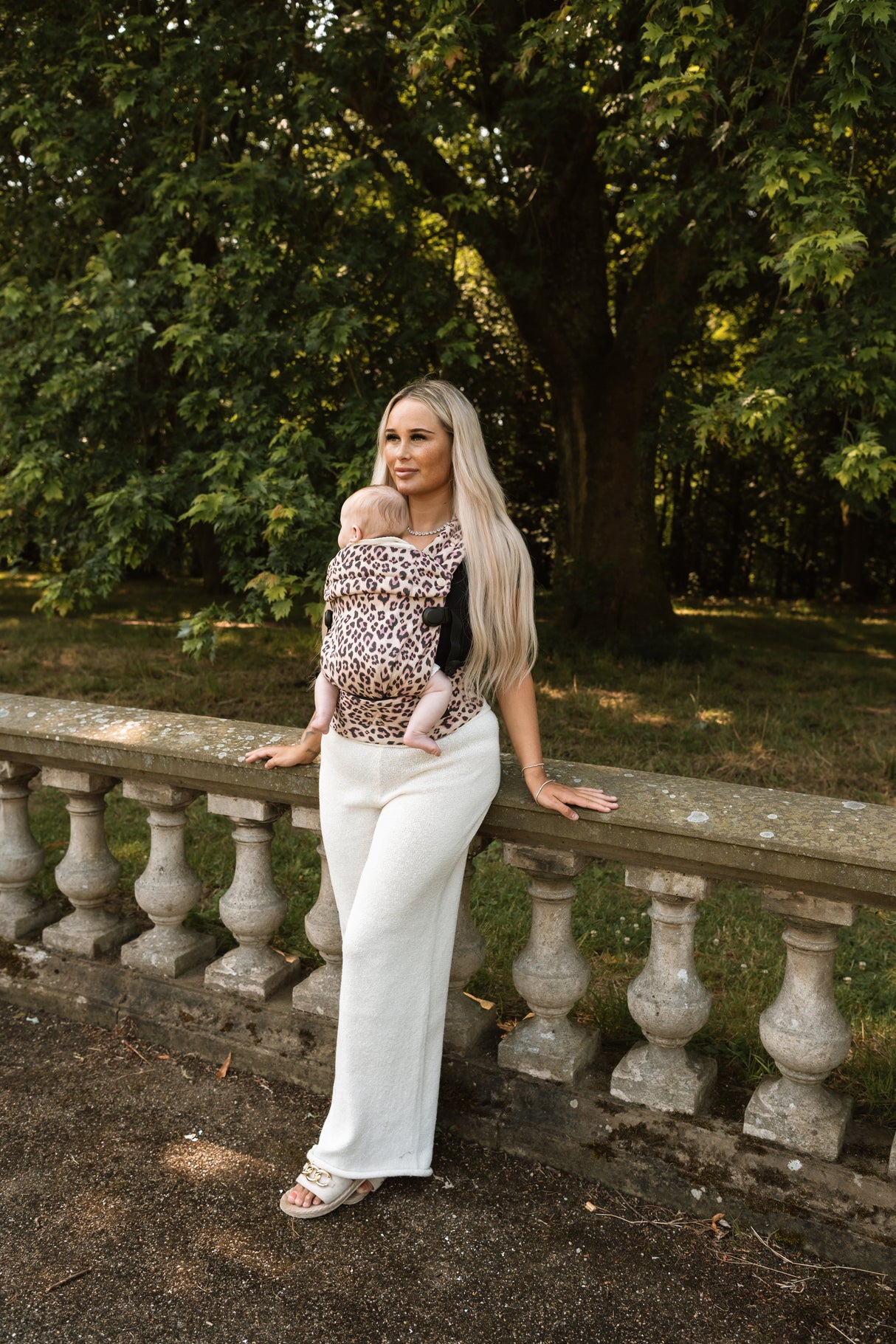 Full body view of woman wearing Nomad Air Dusky Leopard baby carrier leaning on stone balustrade outdoors